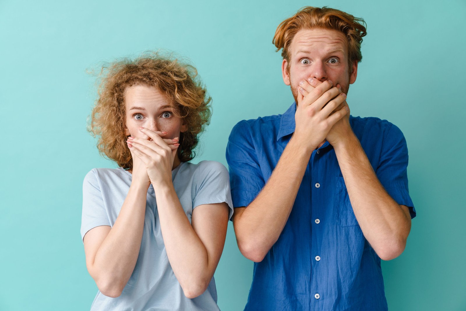 Young surprised redhead couple covering their mouths isolated over blue background